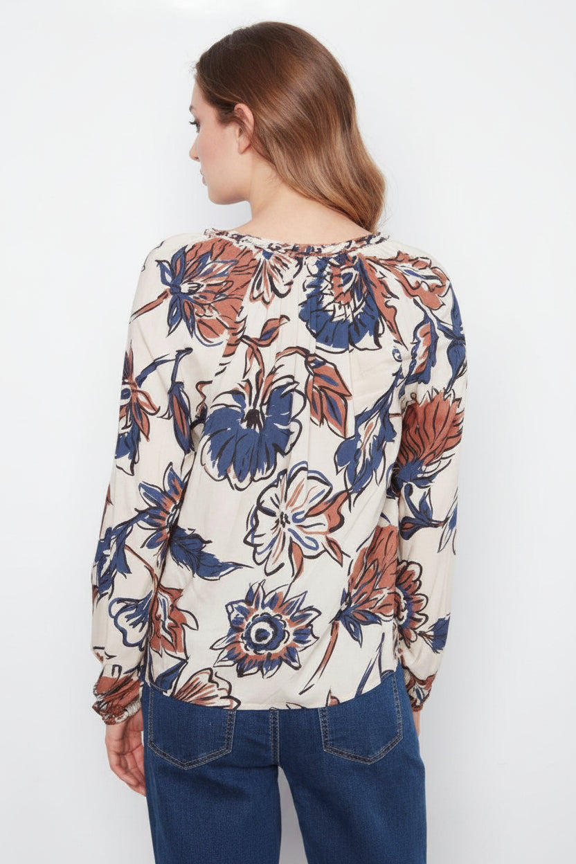 A woman shown from the back wearing a long-sleeve blouse with a cream base and a large floral print in navy blue and rust orange, paired with dark blue jeans against a plain white background.