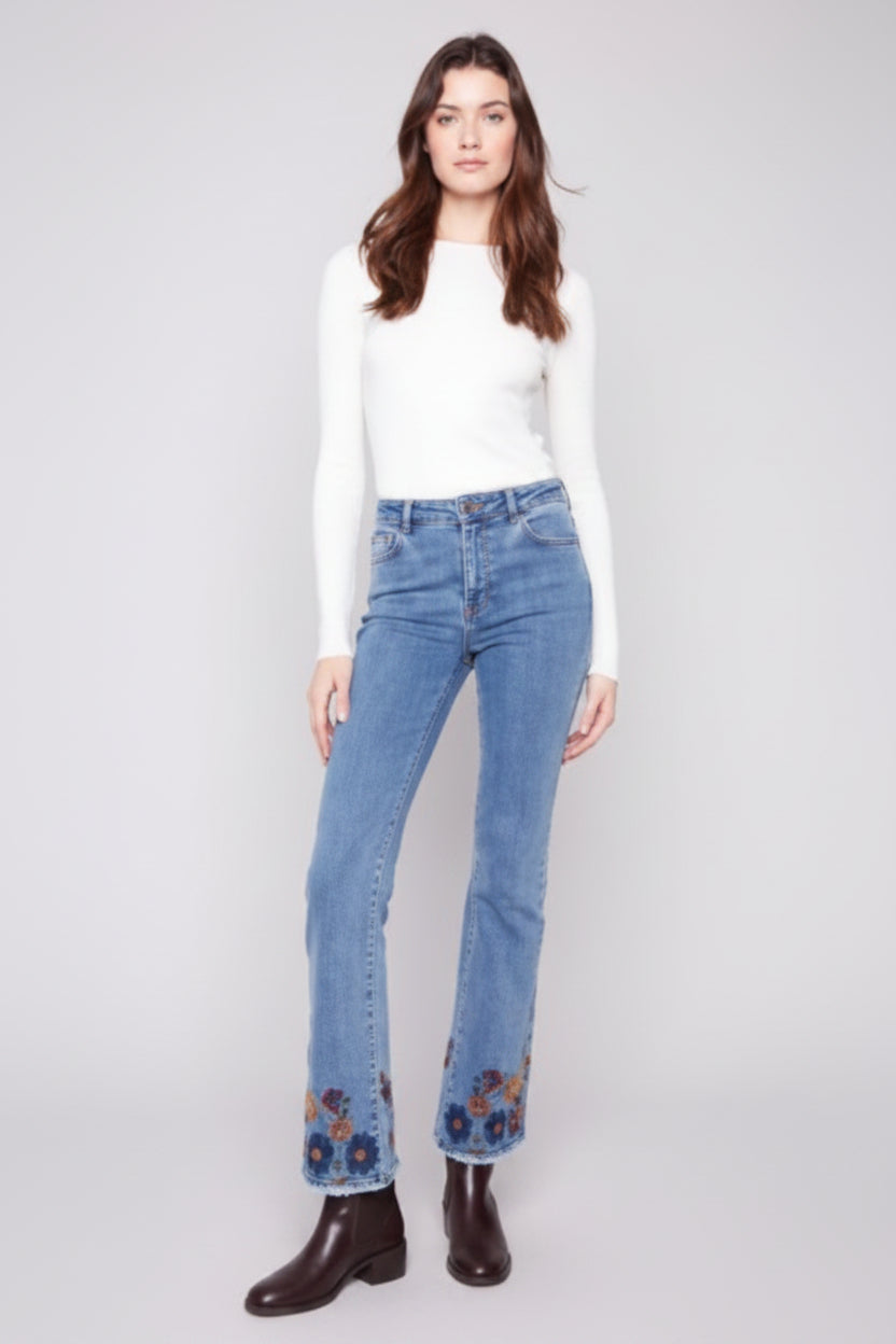 Woman wearing a white top and blue jeans with floral embroidery on a plain background