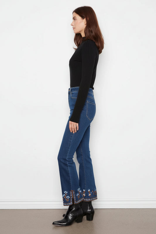 A woman shown in profile wearing a black ribbed long-sleeve top, blue flared jeans with floral embroidery near the hems, and black ankle boots, standing against a plain white wall.
