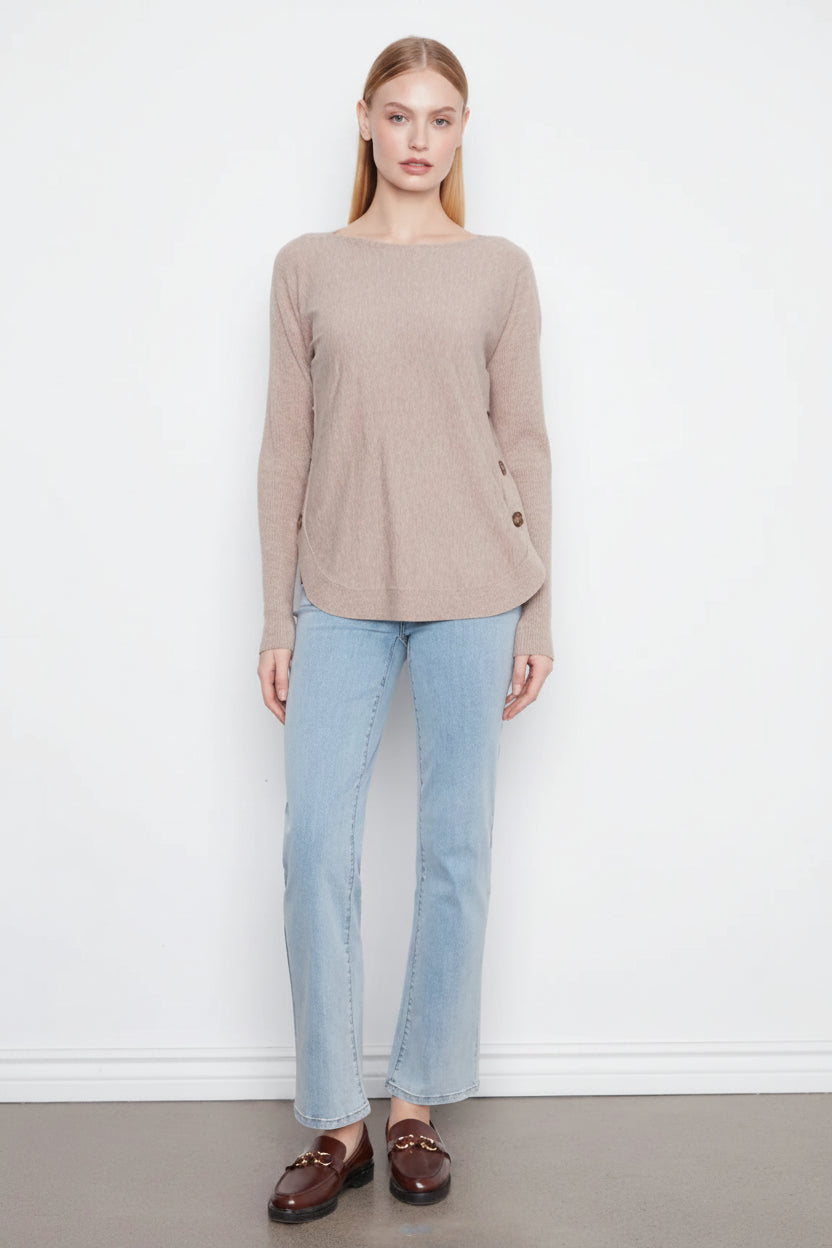 A woman modeling a beige long-sleeve knit top with light blue jeans and brown loafers, standing against a plain white wall.
