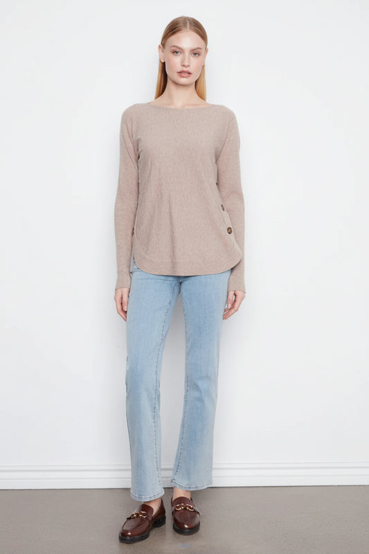 A woman modeling a beige long-sleeve knit top with light blue jeans and brown loafers, standing against a plain white wall.