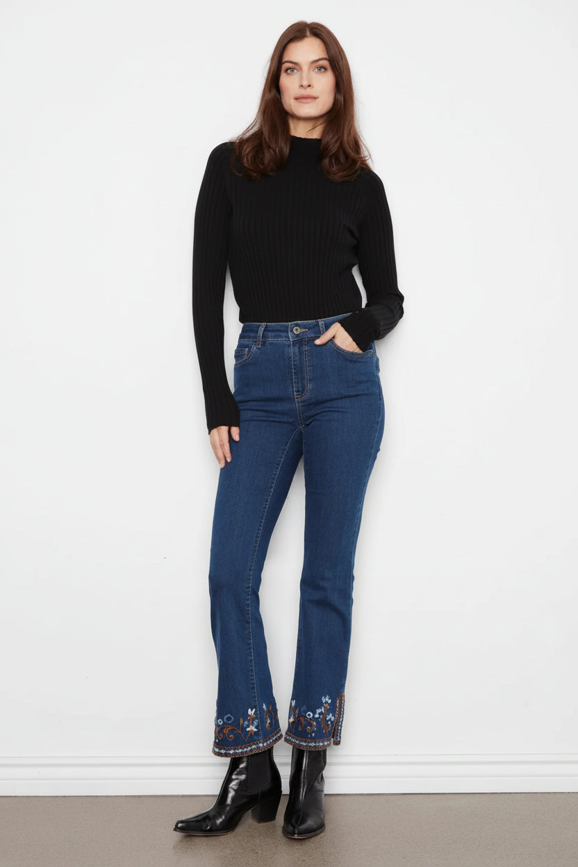 A woman stands against a white wall wearing a black ribbed mock-neck long-sleeve top, blue high-waisted flare jeans with floral embroidery at the hem, and black leather ankle boots.