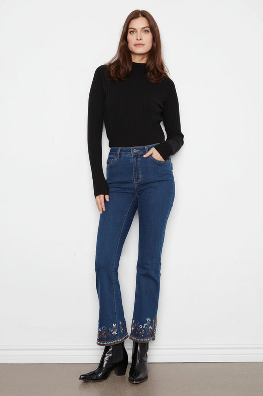 A woman stands against a white wall wearing a black ribbed mock-neck long-sleeve top, blue high-waisted flare jeans with floral embroidery at the hem, and black leather ankle boots.